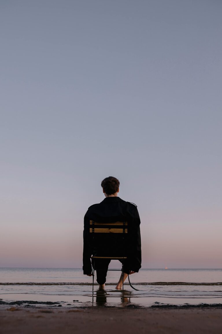 A man sits on a chair facing the calm sea at dusk, creating a tranquil moment.